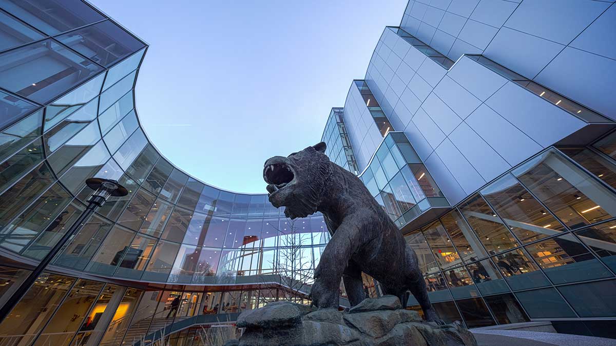 A large bronze tiger statue stands in front of a modern glass building with curved architecture on a university campus.