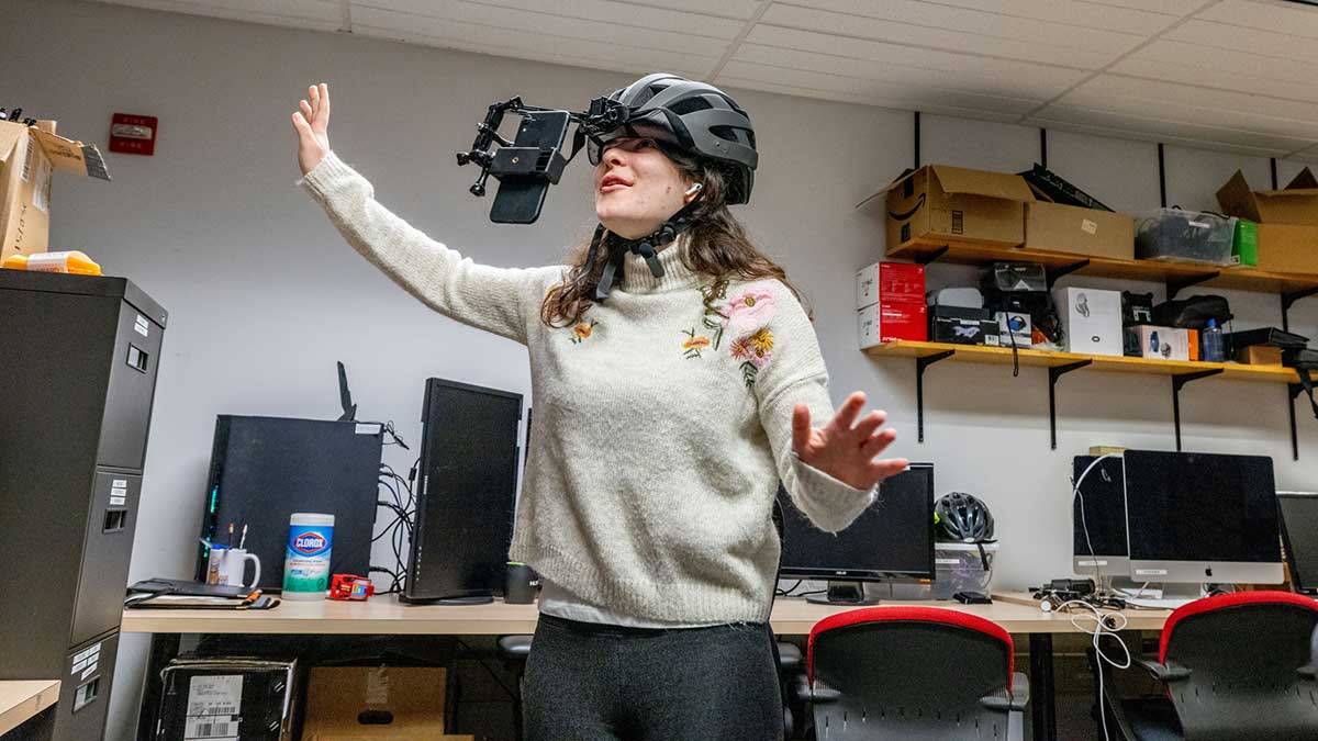 A student wearing a VR headset mounted on a helmet gestures with both hands in a lab filled with computers and technology equipment.