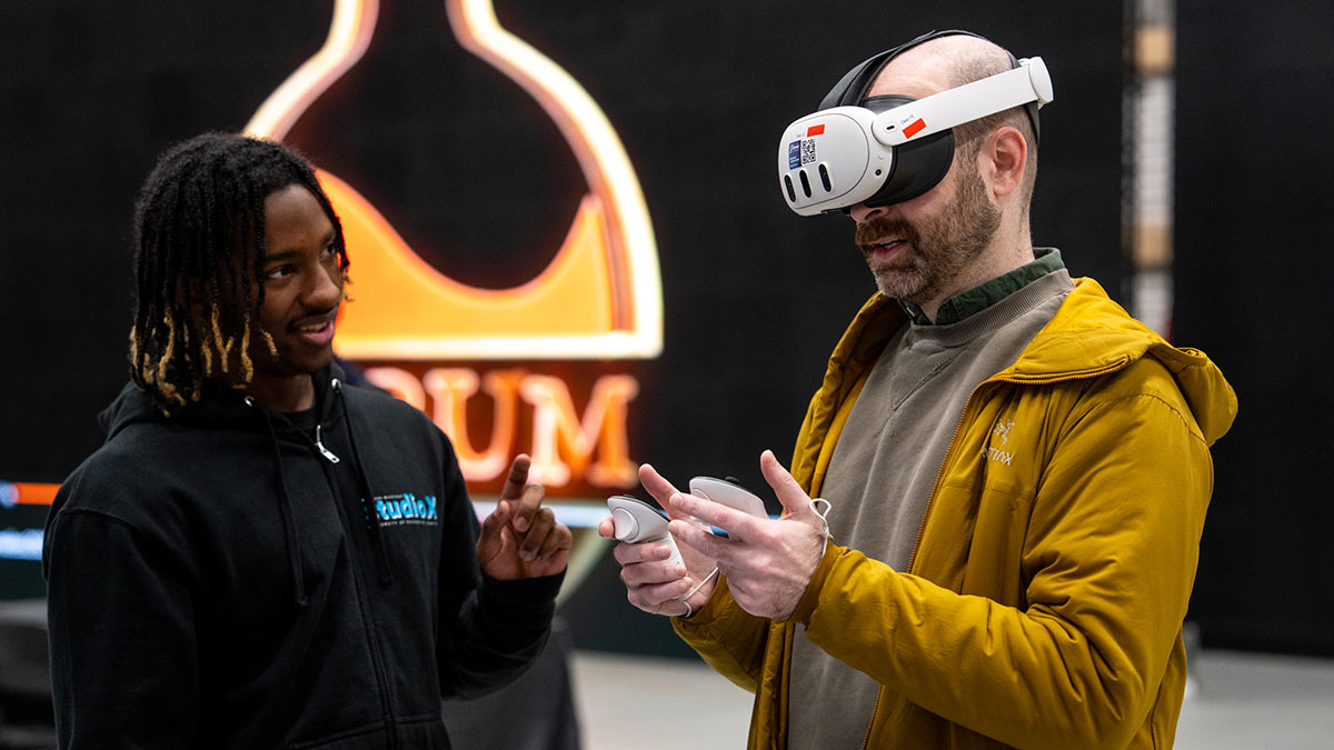 a student talks to a symposium participant who is wearing a virtual reality headset.