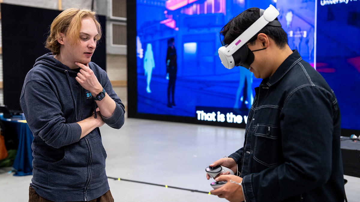 a student talking to a symposium attendee who is wearing a virtual reality headset.