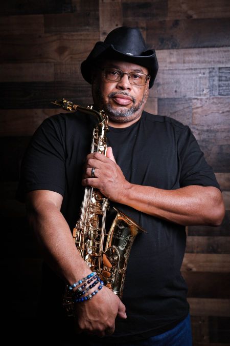 Jimmy Highsmith Jr stands in front of a wood wall holding a saxophone wearing  a black cowboy hat and shirt