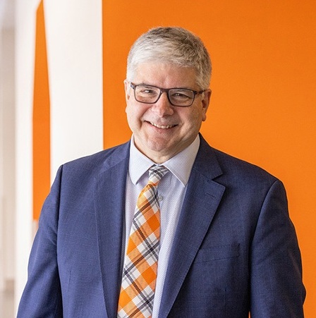 William Sander wearing a blue suit stands with arms crossed in front of an orange wall.