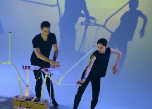 Two people in motion with unique instruments in front of them.