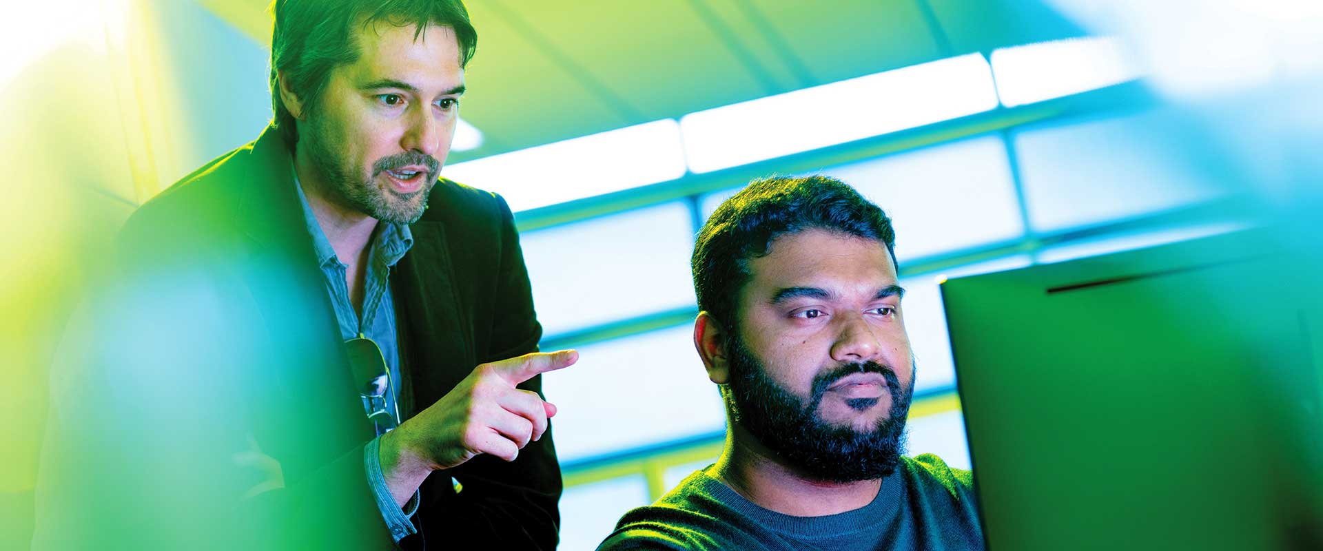 A professor points at a computer screen while a student watches closely in a tech lab.