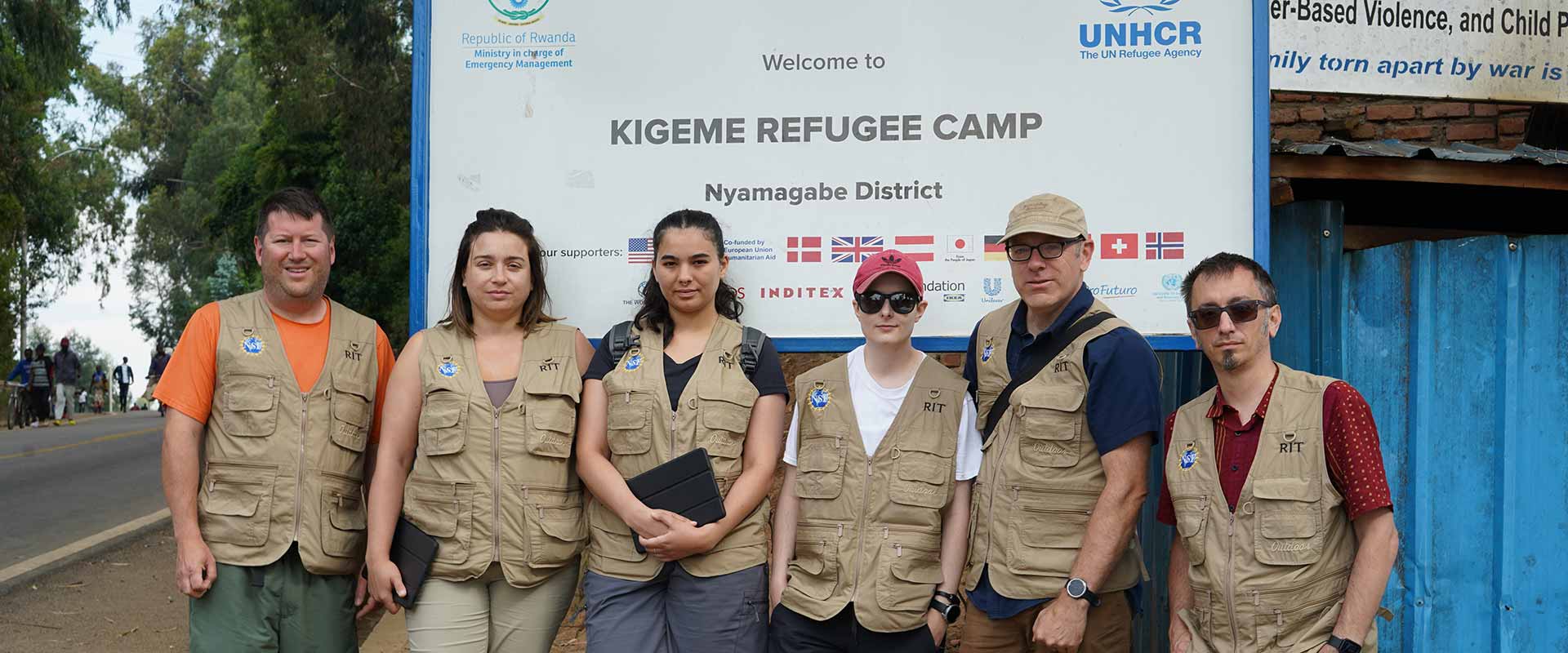 a group of researchers wearing brown field vests standing in front of a sign that reads, Kigeme Refugee Camp.