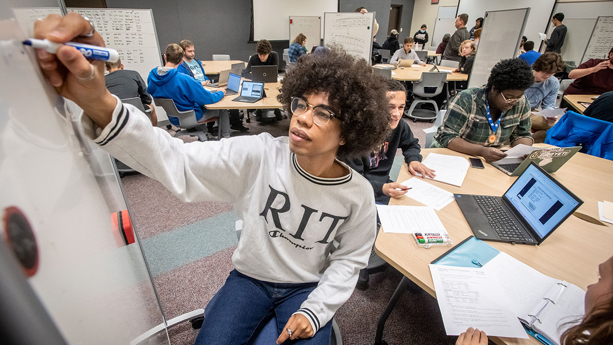 Student in a classroom, writing on a white board.