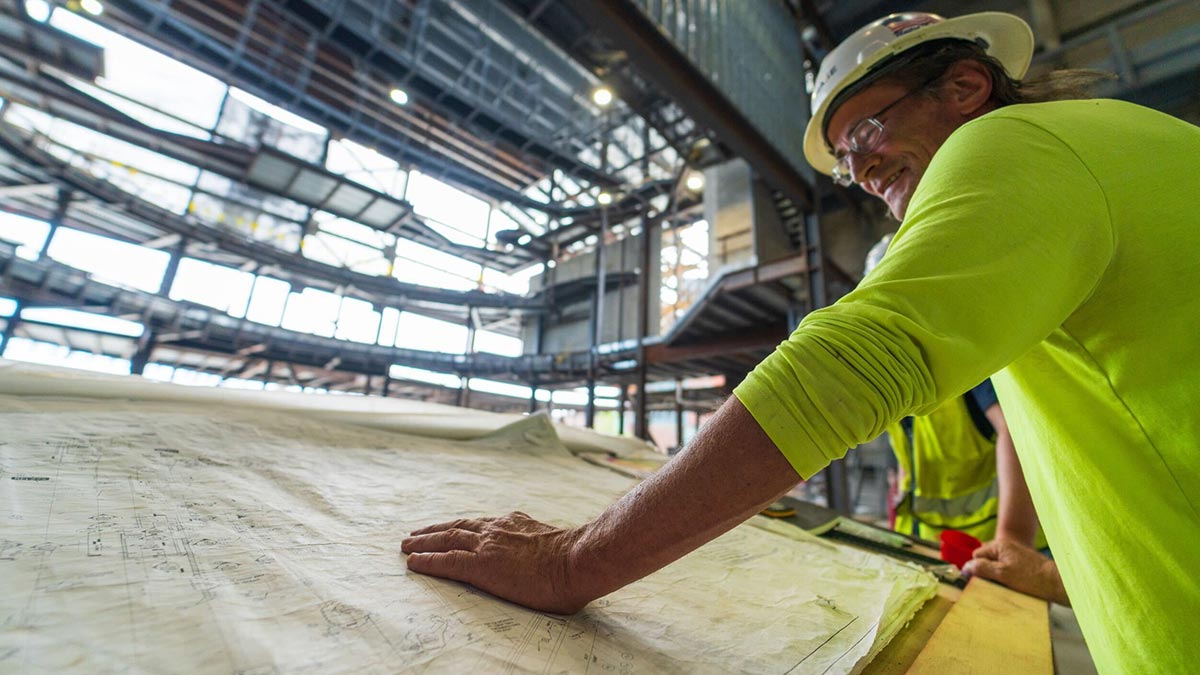 a man wearing a hard hat and high-visibility shirt looks over blueprint plans on a desk while standing in a construction area.