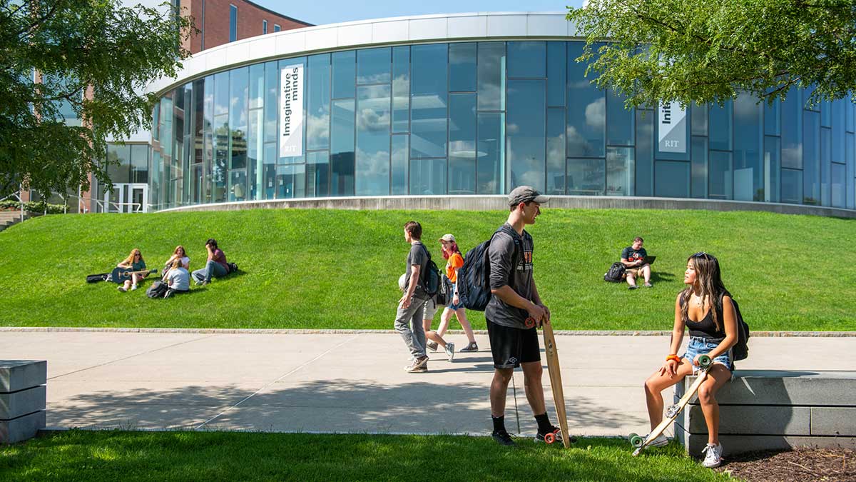 R I T students sit in green grass and walk along a walkway outside of a round, glass building.