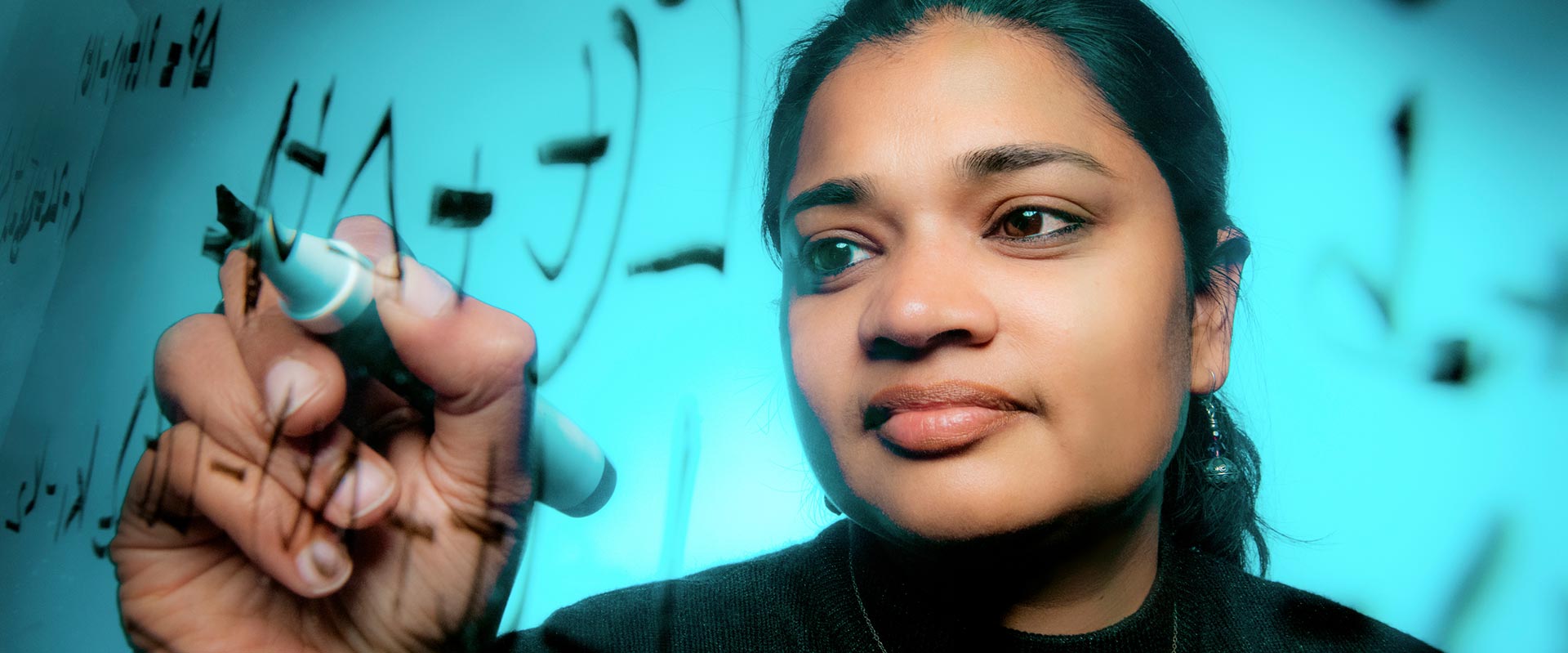 an R I T professor writes math equations on a clear board.