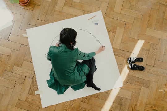 Woman draws semi-circle with black pen on large white paper on a wood floor.