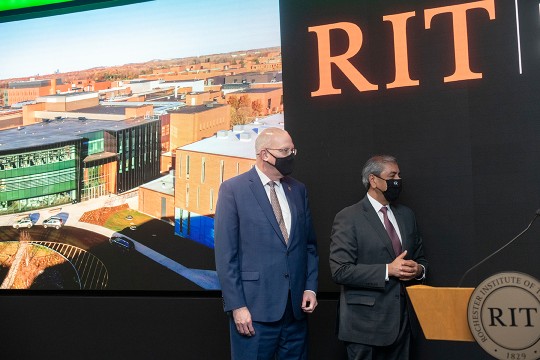 two men standing near a podium in front of a screen displaying a building and the RIT logo.