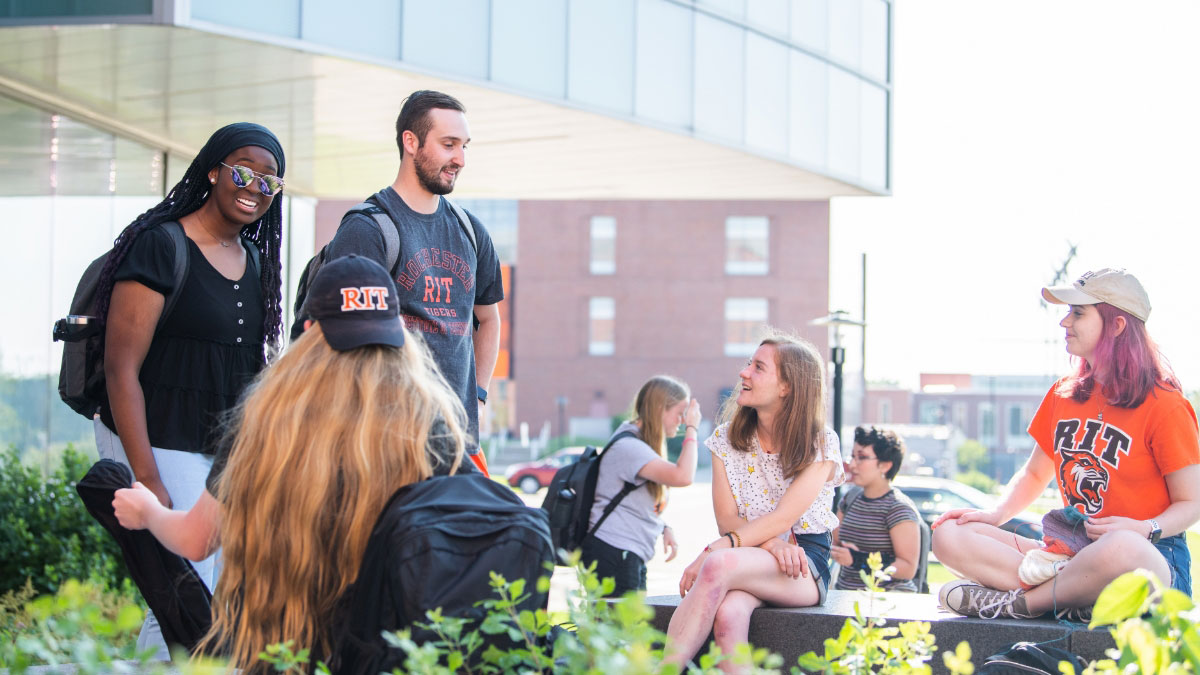 A group of students casually chatting and relaxing outdoors on campus, wearing RIT-branded clothing.