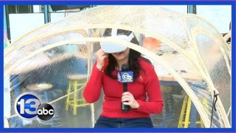A reporter wearing a VR headset holds a 13abc mic inside a transparent dome structure.