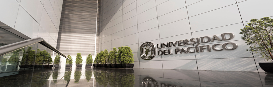 A modern entrance with silver walls, potted trees, and a metallic “Universidad del Pacífico” sign reflected on a polished floor.
