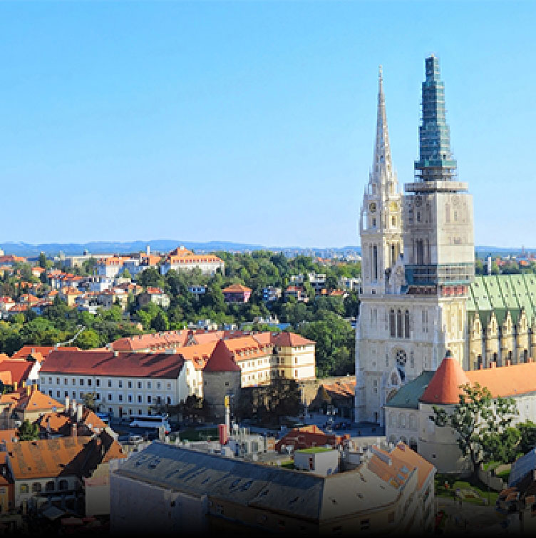 A panoramic view of Zagreb, showcasing the iconic cathedral with its twin spires, surrounded by historic red-roofed buildings and greenery under a bright blue sky.