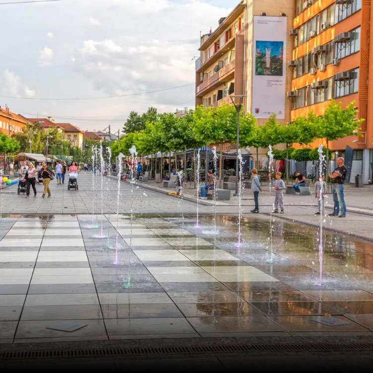A lively street in Pristina with colorful fountains, people walking, outdoor cafes, and vibrant buildings lining the sidewalk.