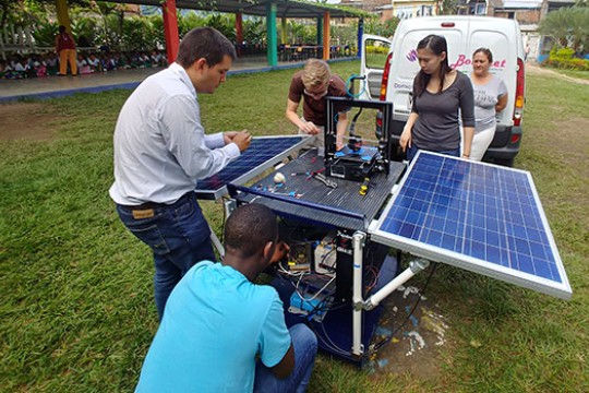RIT students and Professor Rojas are outside setting up their solar powered 3D printer. In the distance, a large group of school children look at their progress.
