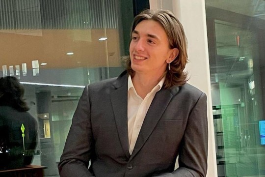 a college age boy is shown in a suit jacket and white shirt leaning on a staircase railing and looking towards the left.