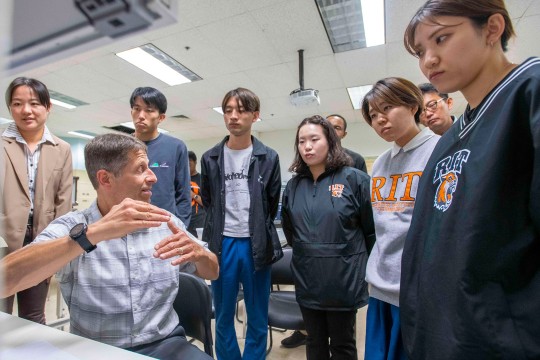 a man sitting at a computer turns his back to the monitor to speak to seven japanese students in a classroom