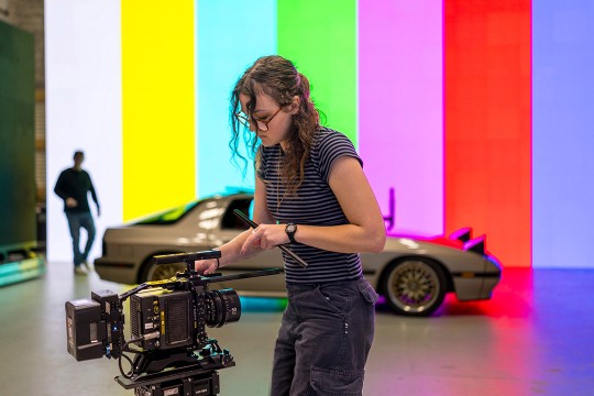a student operates a camera in front of a multicolor screen with a car sitting in front of it