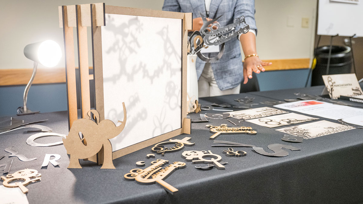 A person demonstrates a shadow puppet display with various cutout shapes on a table under a lamp.