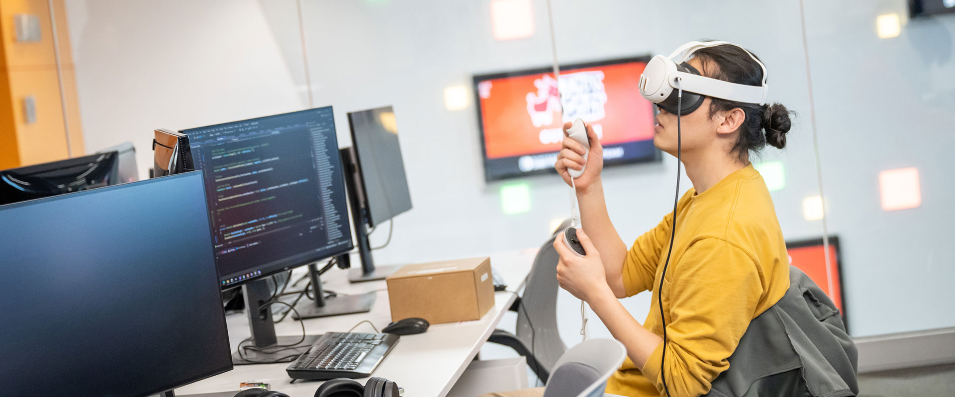 A student wearing a VR headset interacts with a controller while sitting at a desk with multiple monitors.