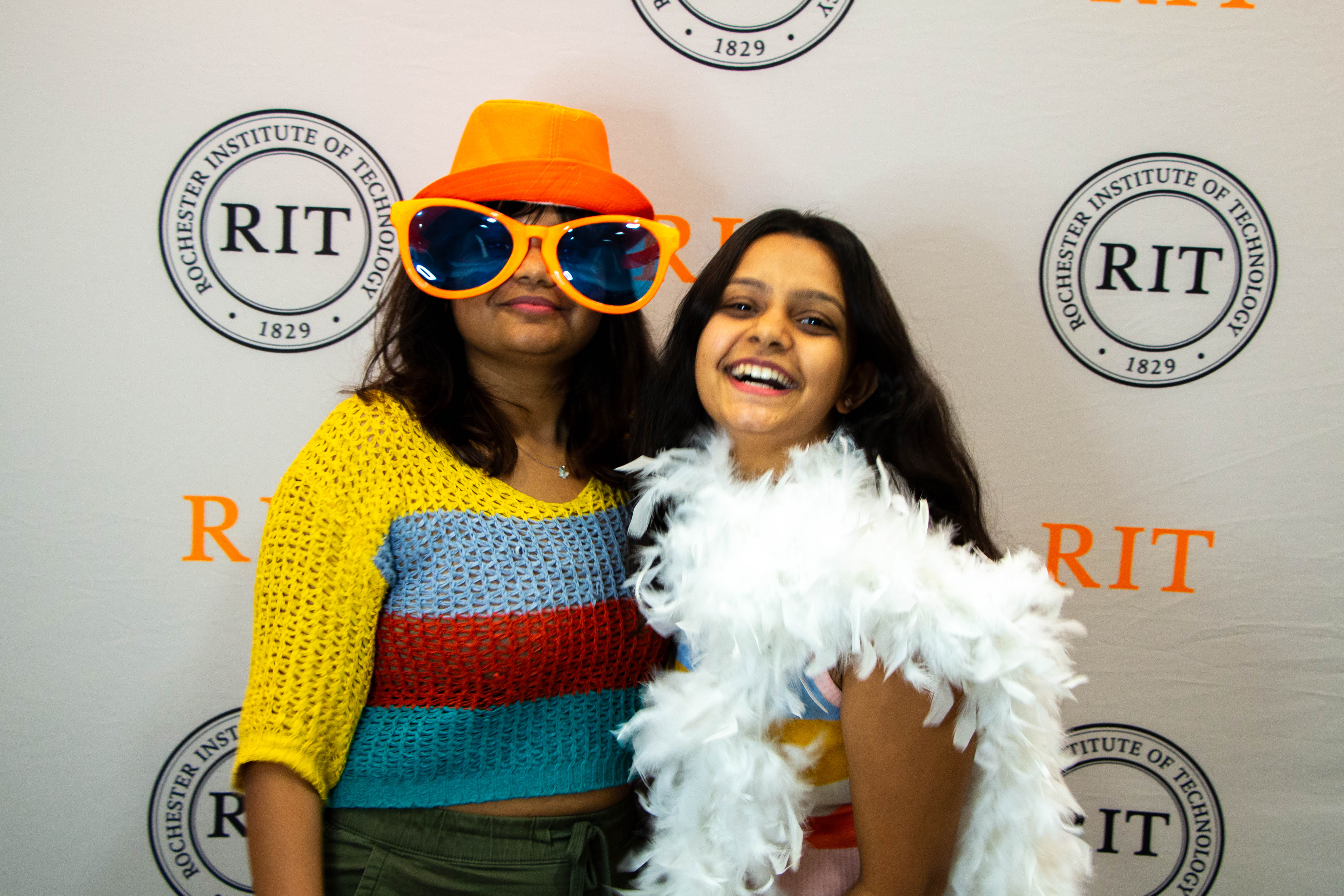 two students in front of a RIT logo background in orange hats and googles as props of the photobooth