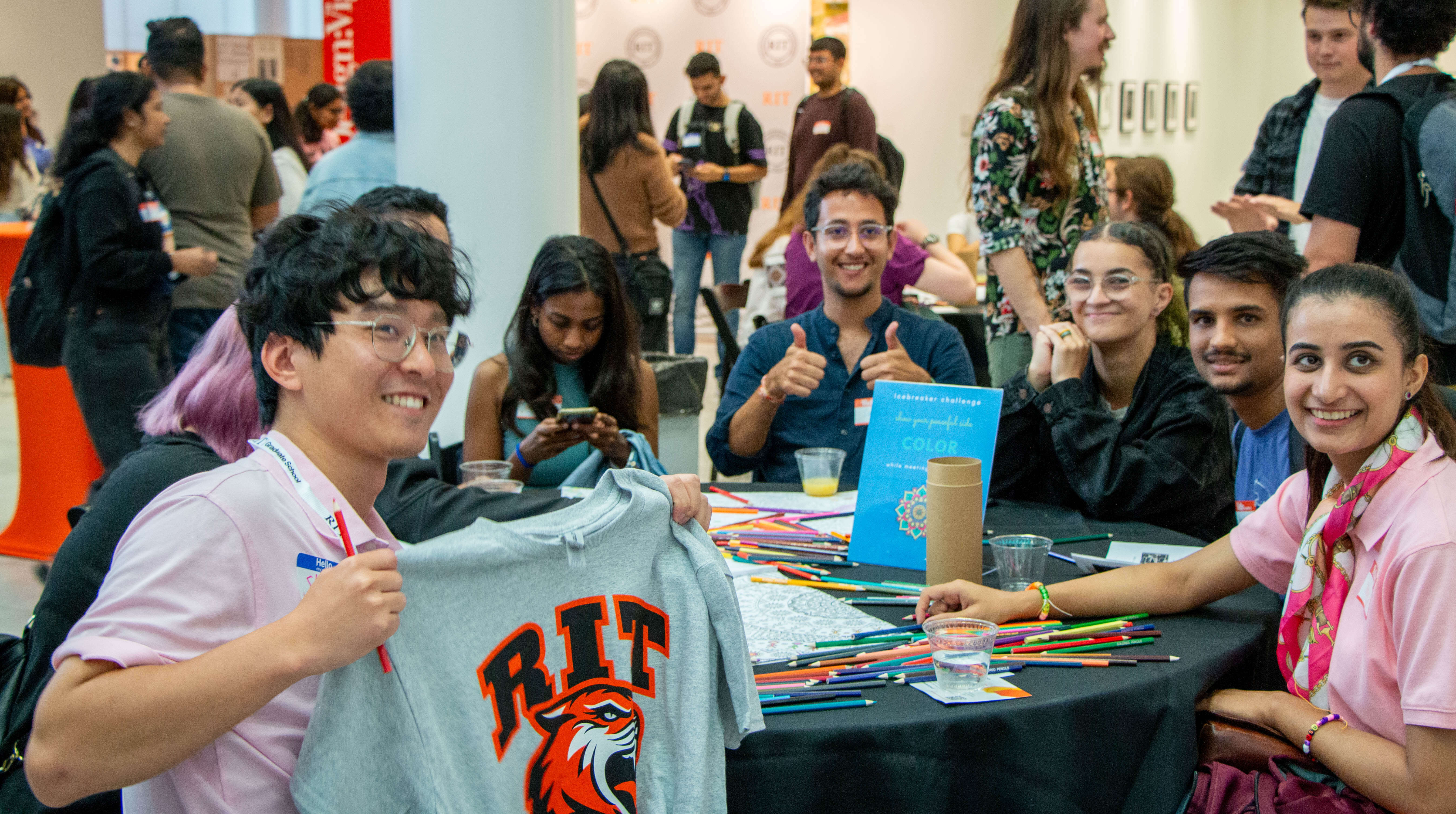 students pose for a picture on a circular table with one student posing with their RIT tshirt