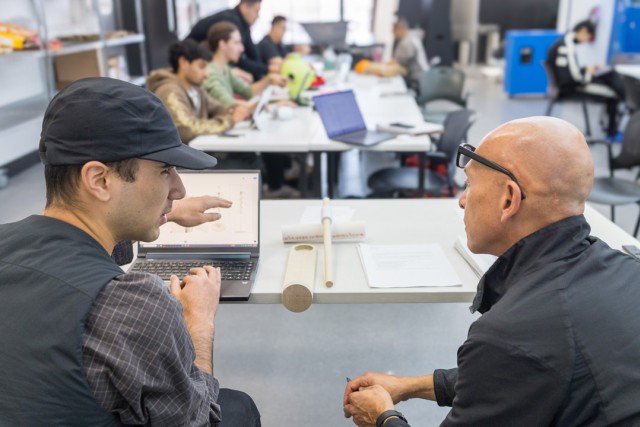 a student in a black hat speaks with a man sitting to his right, gesturing near a laptop screen.
