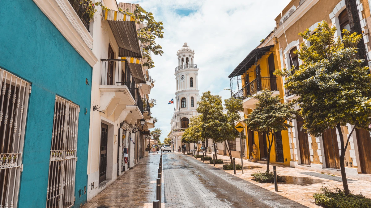 A quiet, sunlit street with wet paving stones runs between turquoise and yellow colonial buildings with balconies and trees, leading toward a white clock tower with a flag in the distance.