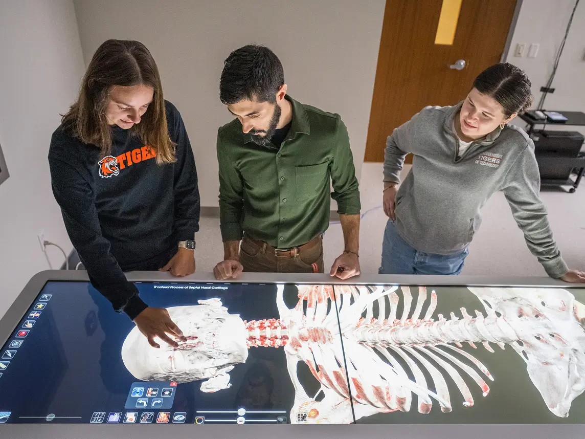 Three people standing at the anatomy table looking at a digital skeleton.