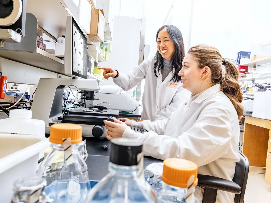 Two people in lab coats, looking at a display showing what is under the microscope.