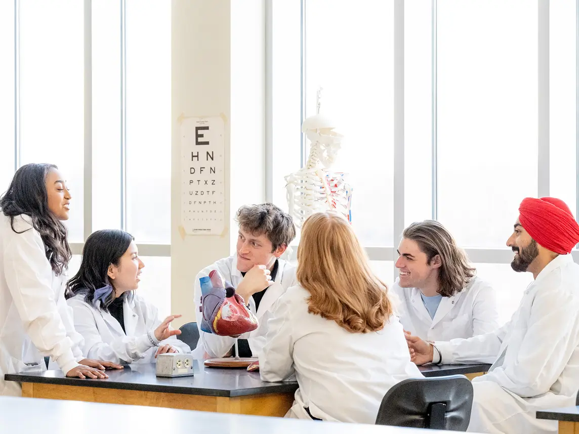A group of six students in lab coats sitting around a table having a conversation.