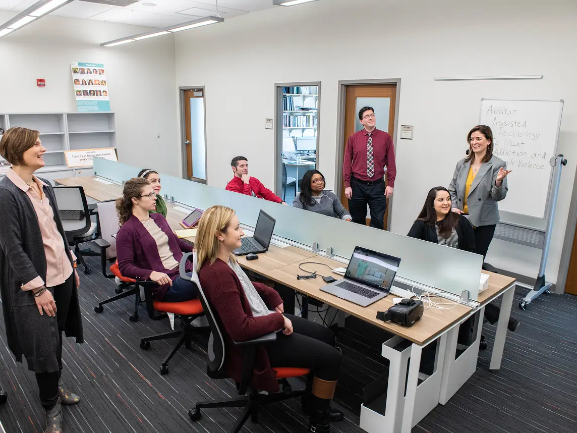 Six students sitting at desks with three instructors standing around them.