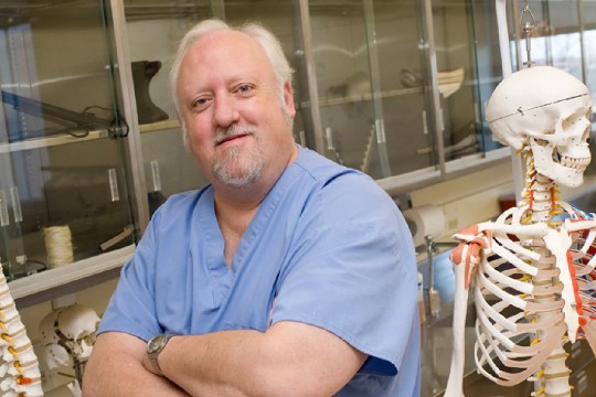 a man with white hair stands in a lab next to skeletons.