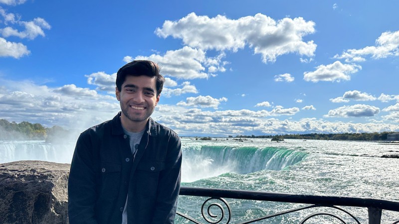 RIT student Sammy Doel wears a black jacket and stands in front of a railing overlooking Niagara Falls.