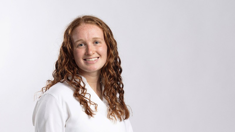 RIT student Madison Degenfelder has long, curly brown hair and is wearing a plain white top, posed against a neutral light gray background.