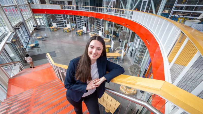 A young woman in business attire smiles at the camera while leaning on a railing at the top of a bright orange staircase inside a modern, multi-level building with glass walls, open seating areas, and a spacious atrium below.