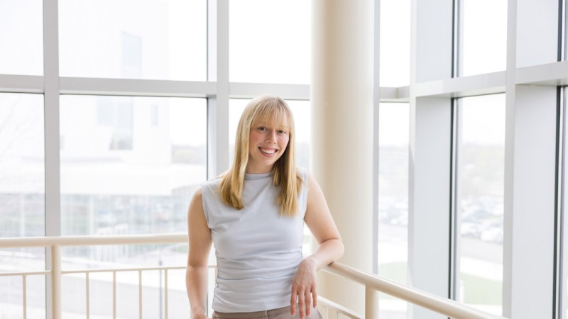 A portrait of RIT student Jillian Antol, who is wearing a neutral-colored top and gold necklace, smiling in a brightly lit, modern glass atrium on campus.