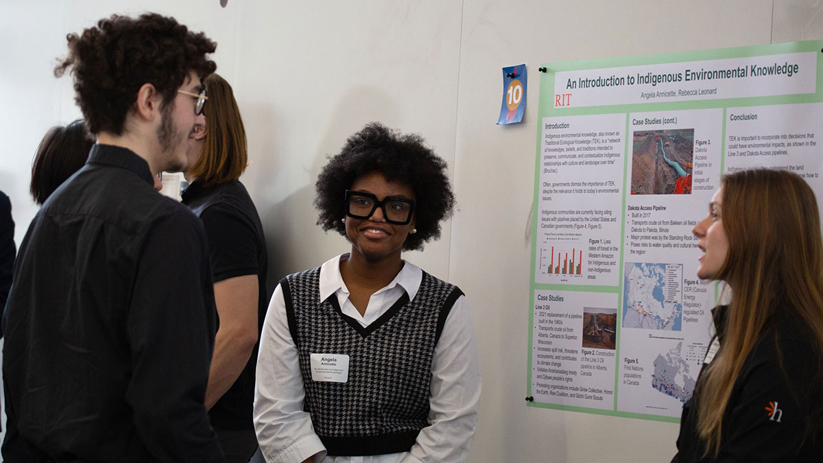 A student presenter stands beside a poster on Indigenous environmental knowledge while speaking with attendees at a symposium.