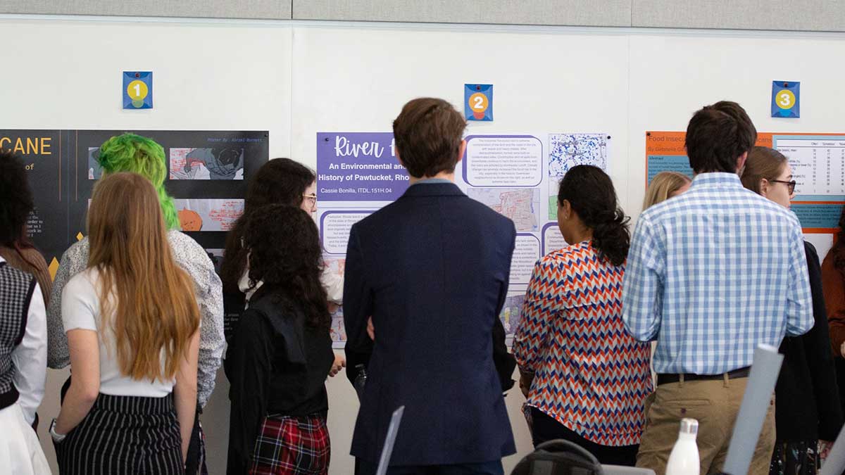 Attendees gather in front of multiple research posters at a student symposium.
