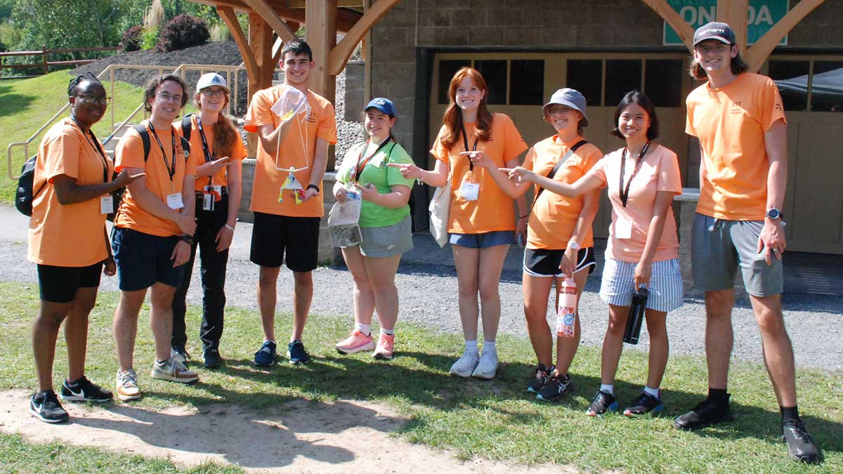 A group of students in orange shirts stands outside smiling and pointing to a small object held by one team member.