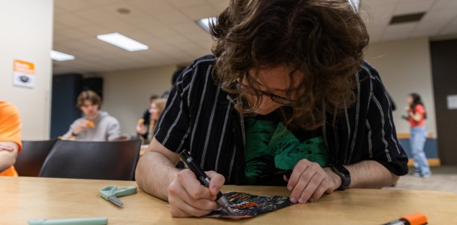 Honors Student wearing a black shirt with white vertical stripes, and curly brown hair working on a 6x6 piece of art with a sharpie. Other blurred Honors students in the background.