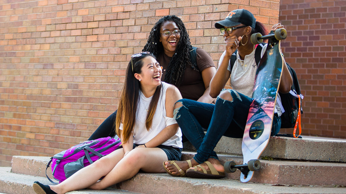 three students sit in front of a brick building, one holds a longboard