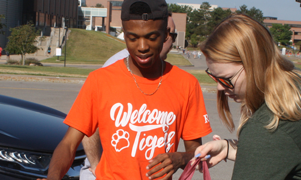 2 students standing next to cars packing bags. Person in white hat loading trunk behind them.