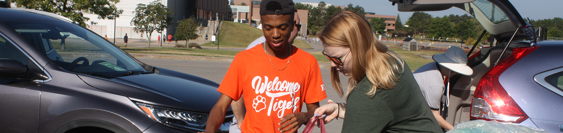 2 students standing next to cars packing bags. Person in white hat loading trunk behind them.
