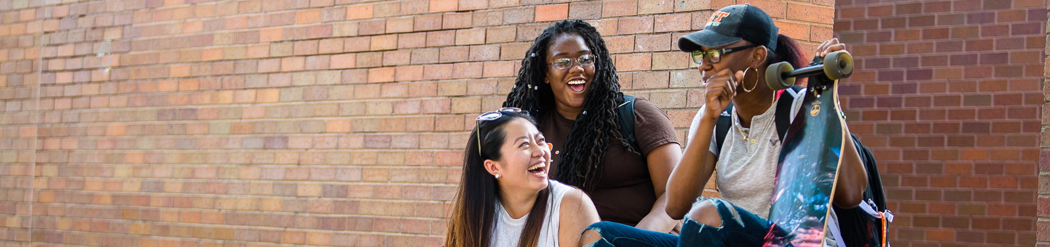 three students sit in front of a brick building, one holds a longboard