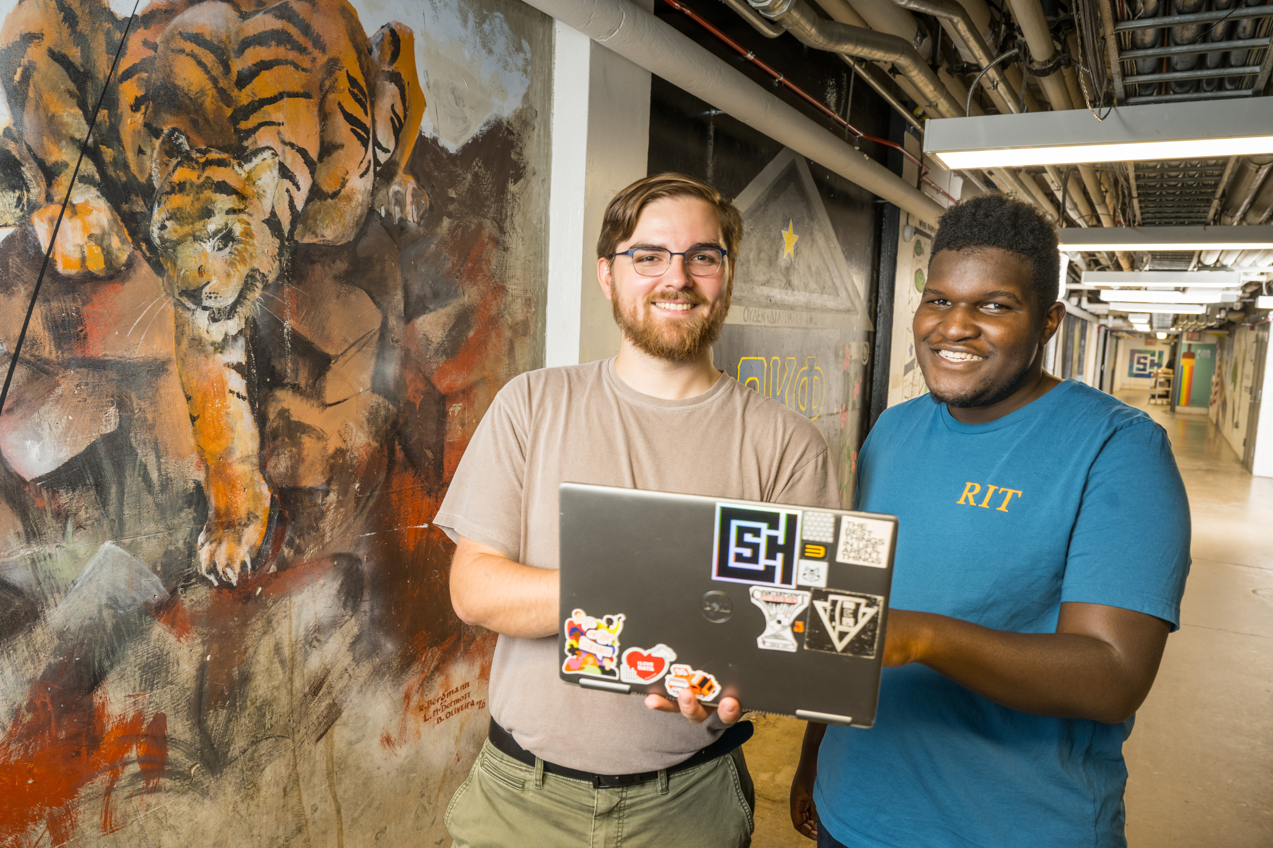 two students smiling at the camera on a laptop next to a mural in the tunnels