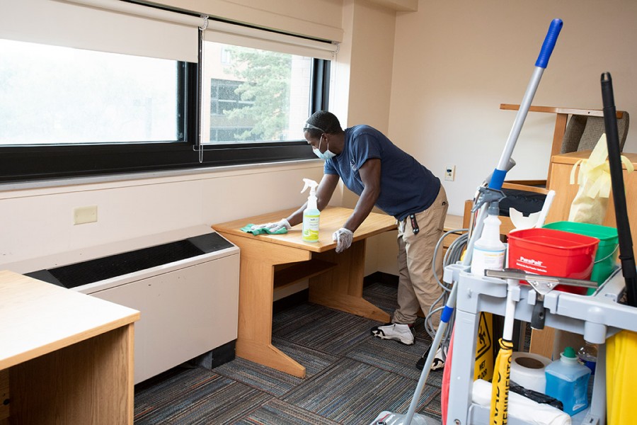 a man wearing a blue shirt wipes down a desk in a residence hall room