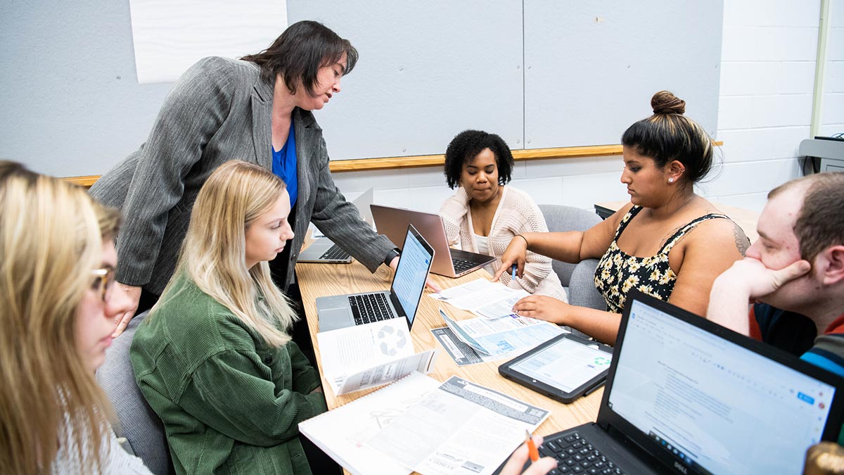 a professor works with students at a table covered with papers and laptops.